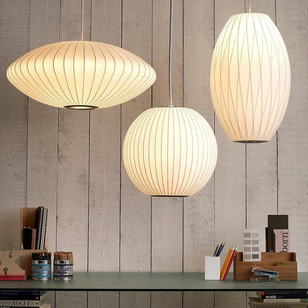 Three lit Clérachic pendants in different shapes, a wide disc, a ball and a tall oval, glow warm white above a shelf of books and pots.