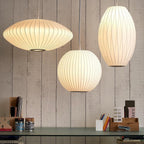 Three lit Clérachic pendants in different shapes, a wide disc, a ball and a tall oval, glow warm white above a shelf of books and pots.