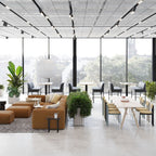A round white Clérachic pendant hangs near the glass wall of a large, light lounge with brown sofas and long tables.