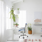 A small round white Clérachic pendant hangs by a window above a white desk, with a trailing fern and a blue office chair.