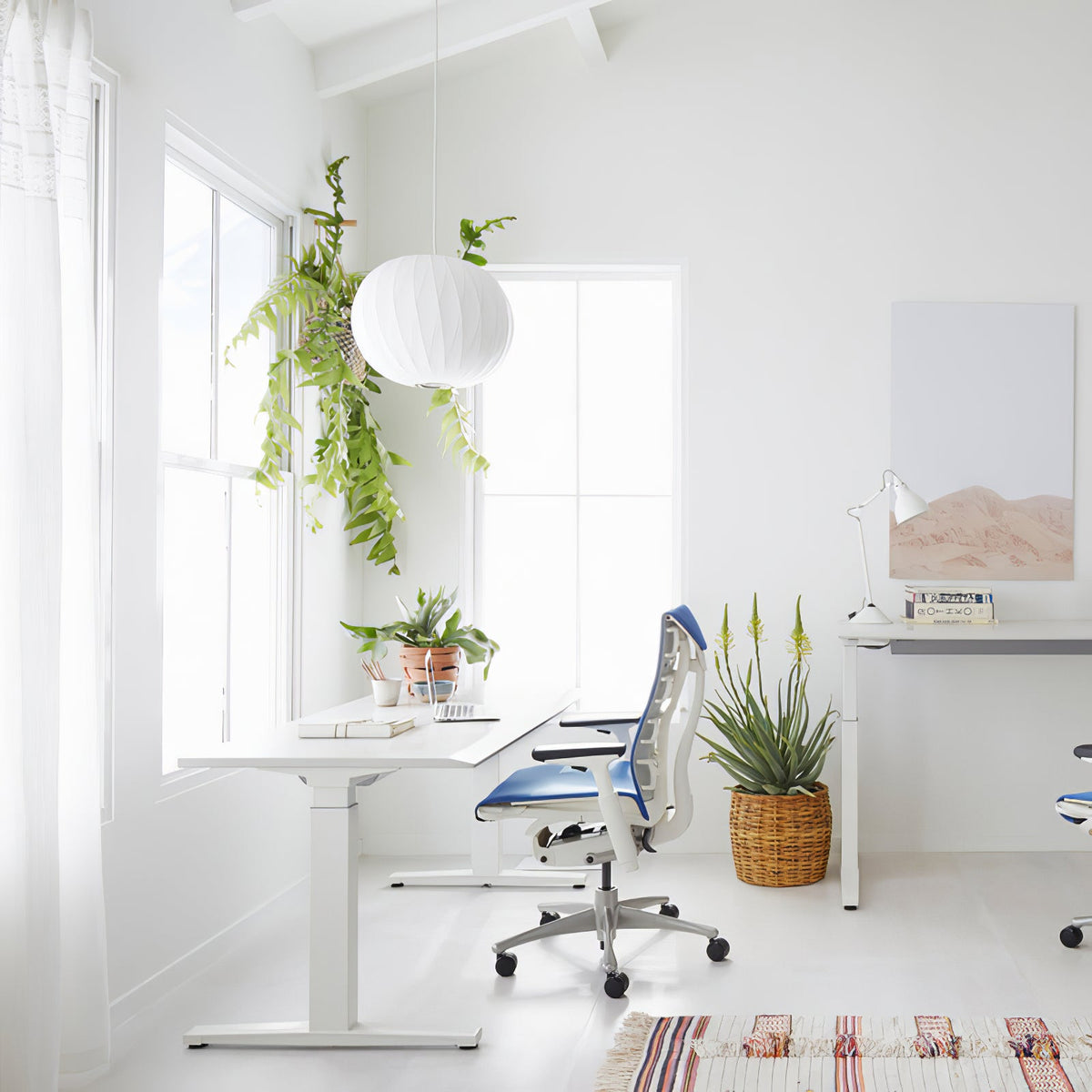 A small round white Clérachic pendant hangs by a window above a white desk, with a trailing fern and a blue office chair.