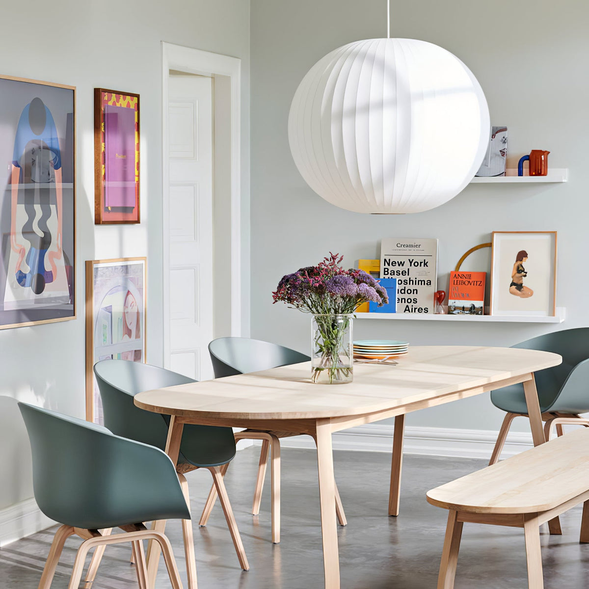 A large round white Clérachic pendant hangs over a long pale wooden table with green-grey chairs, in a room hung with colourful prints.