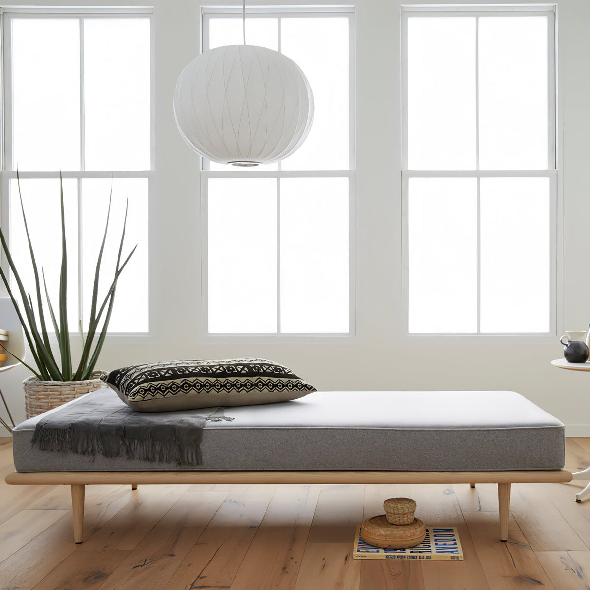 A rounded white Clérachic pendant with a ribbed shade hangs in a bright room above a grey daybed in front of tall windows.