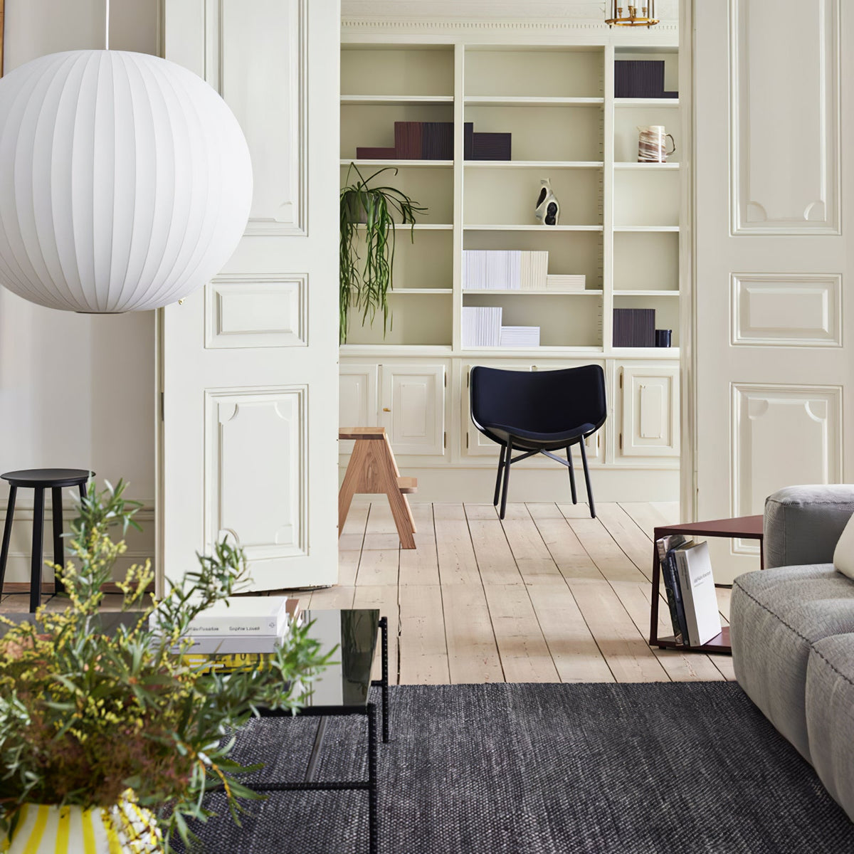 A large round Clérachic pendant with a pale ribbed shade hangs by a white door in a room with a dark chair and a tall bookcase.