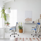A small round white Clérachic pendant hangs above a bright white desk, near a hanging plant and a blue mesh chair.