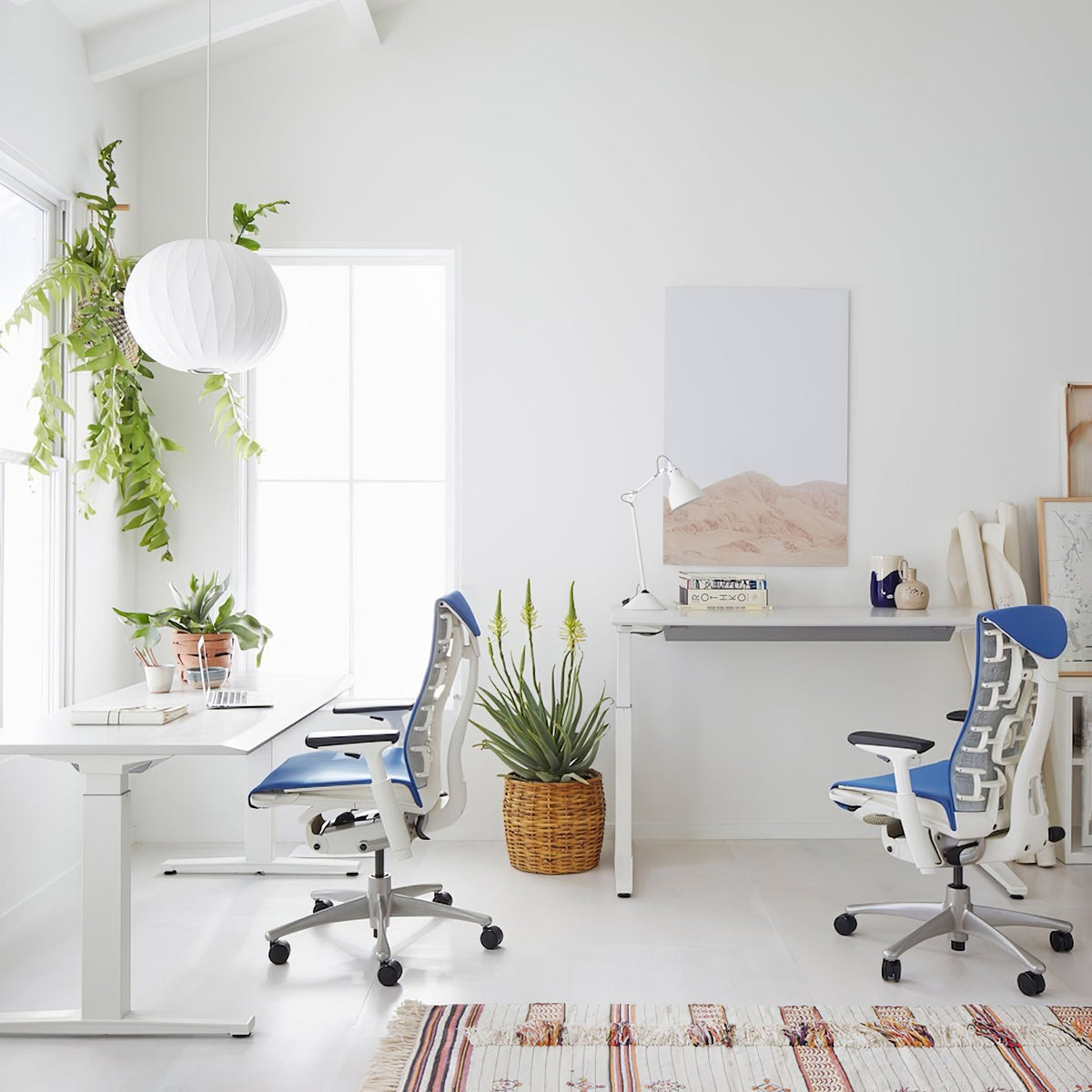 A small round white Clérachic pendant hangs above a bright white desk, near a hanging plant and a blue mesh chair.