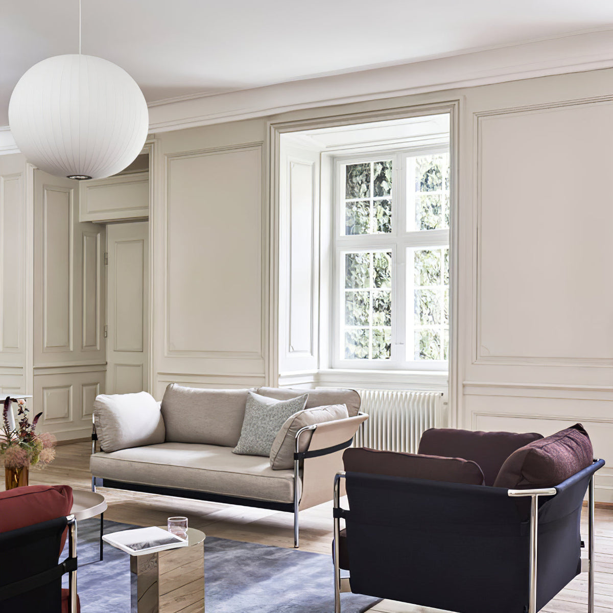 In a panelled living room, a large round white Clérachic pendant hangs at the top of the frame above a sofa and dark armchairs.