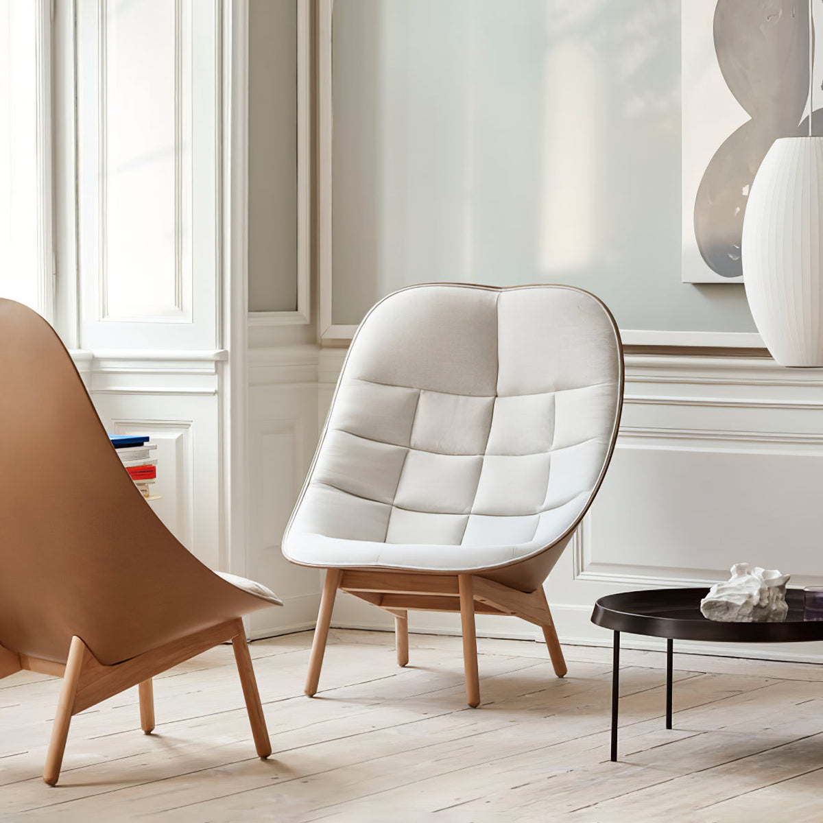 A tall white Clérachic pendant appears at the edge of a pale panelled room with a cream armchair and a low black table.