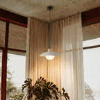 Clérachic white pendant light glowing beside sheer curtains and a large monstera plant near a corner window.