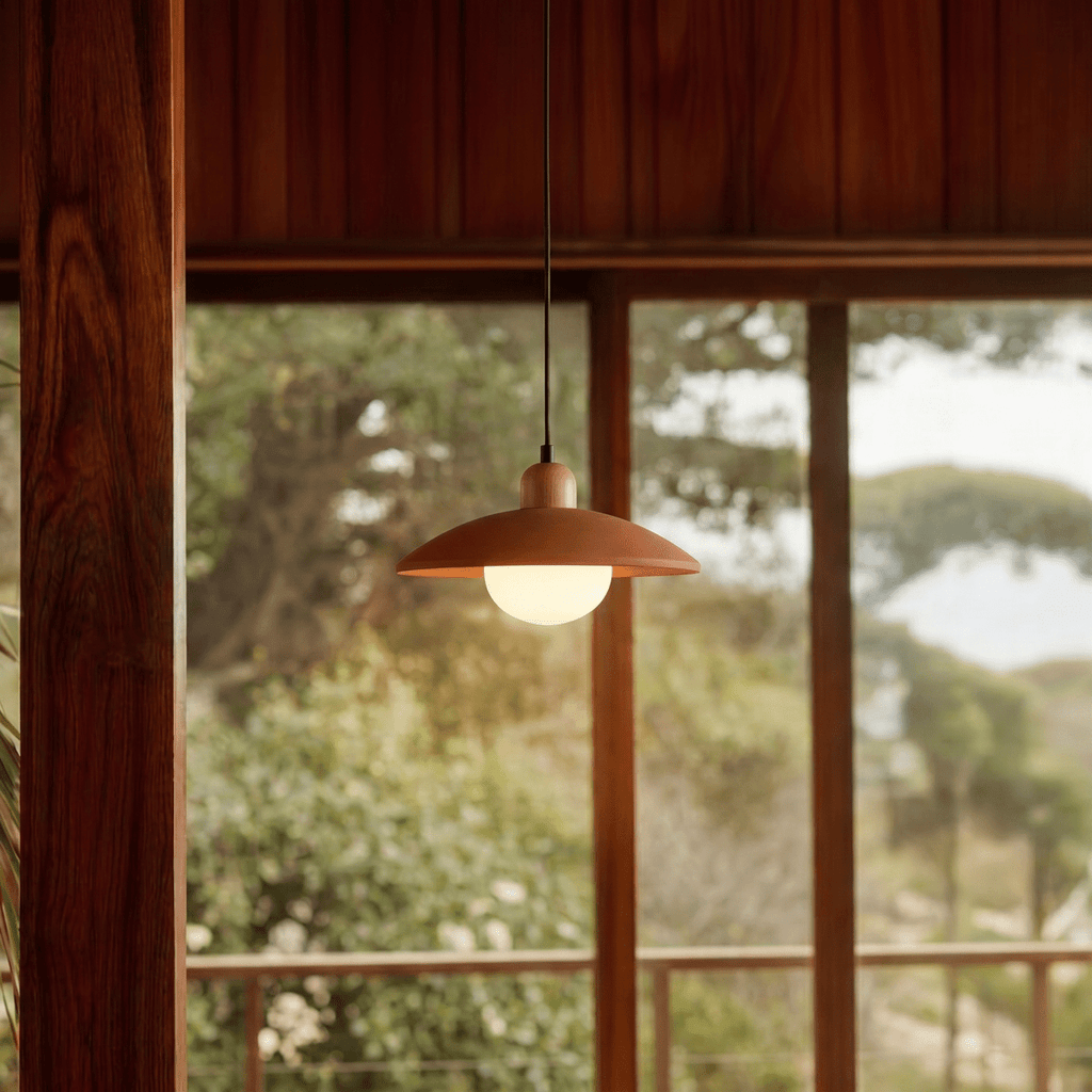 Clérachic orange pendant light glowing beside timber-framed windows overlooking a wooded coastal hillside.
