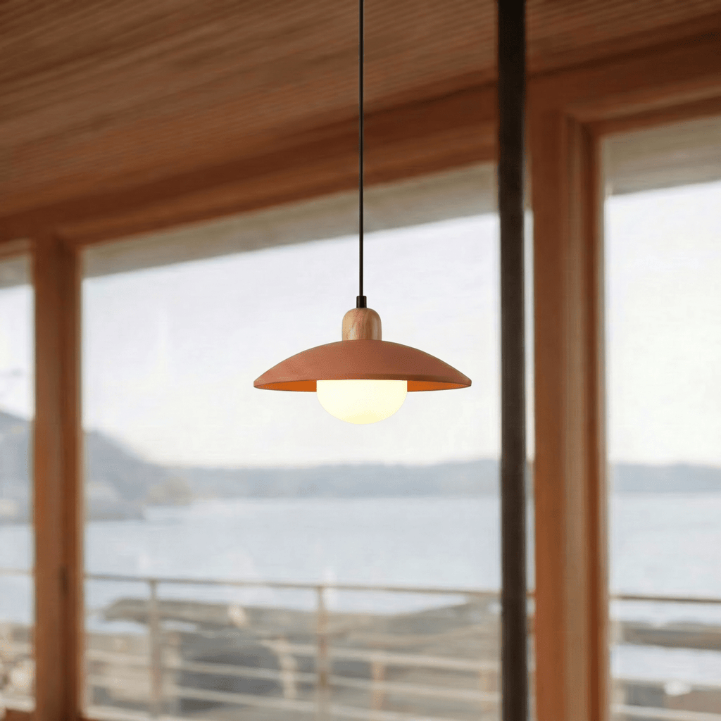 Clérachic orange pendant light glowing beside a window overlooking a calm bay beneath timber ceiling beams.