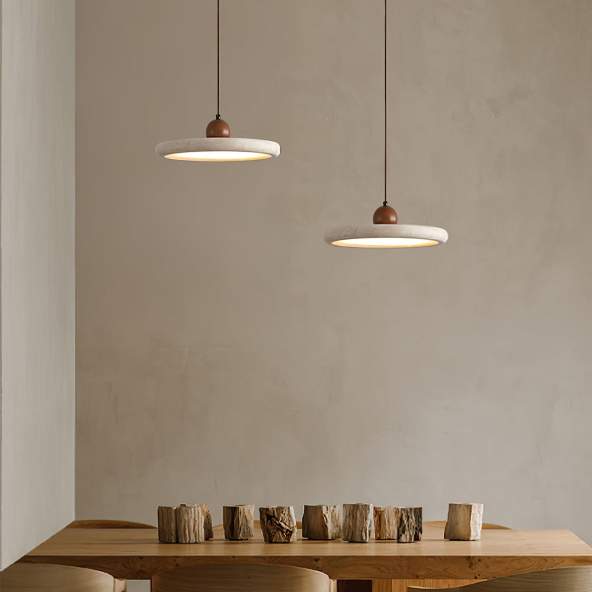 Two Clérachic disc pendants hang at different heights over a wooden table with a row of chopped log pieces, against a plaster wall.