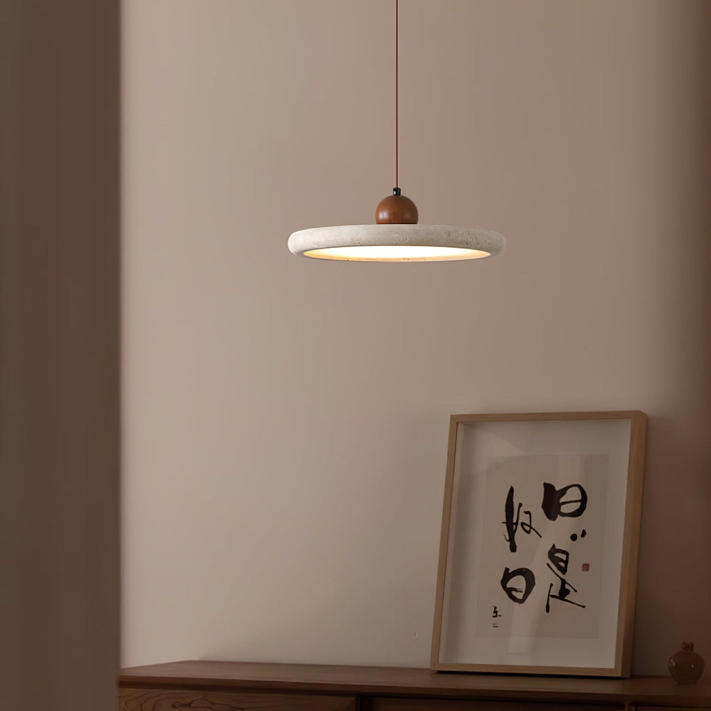 A lit Clérachic disc pendant with a walnut bead hangs against a dusty pink wall, beside a framed ink calligraphy print on a sideboard.