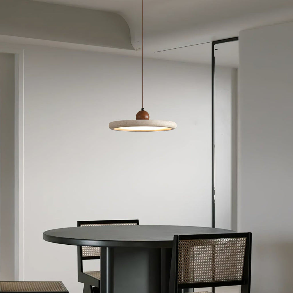A Clérachic travertine-look disc pendant hangs over a round dark dining table with cane-backed chairs, in a pale, minimal room.