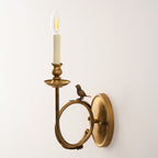 Clérachic wall sconce in warm antique brass, its candle-style bulb glowing softly against a plain cream wall.
