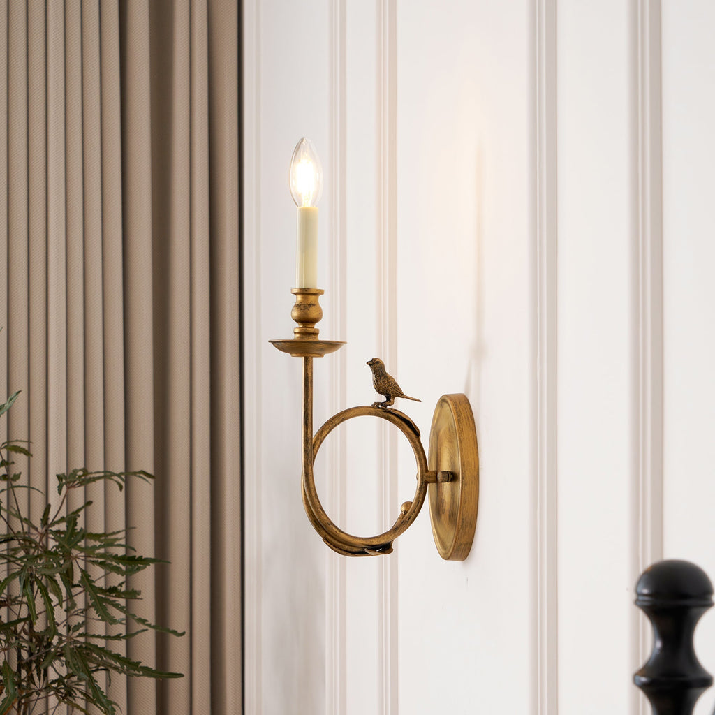 The Clérachic sconce lit beside a curtain and a leafy plant, casting warm light in a styled bedroom corner.