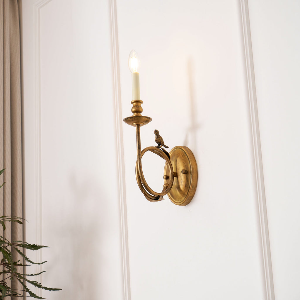 Clérachic's sconce glowing softly on the wall, with green plant fronds framing the lower corner of the shot.