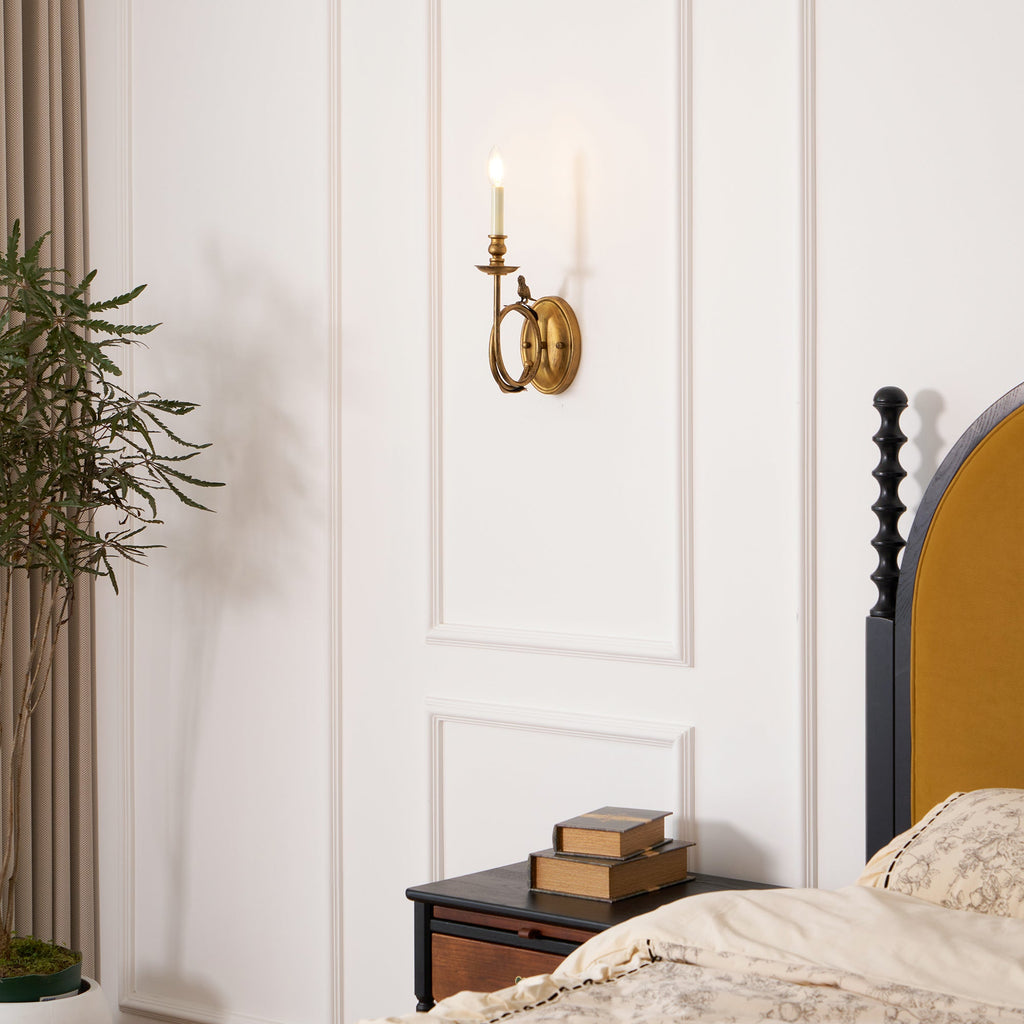 The Clérachic sconce lit beside a dark wood bed frame, with a small nightstand and plant visible nearby.