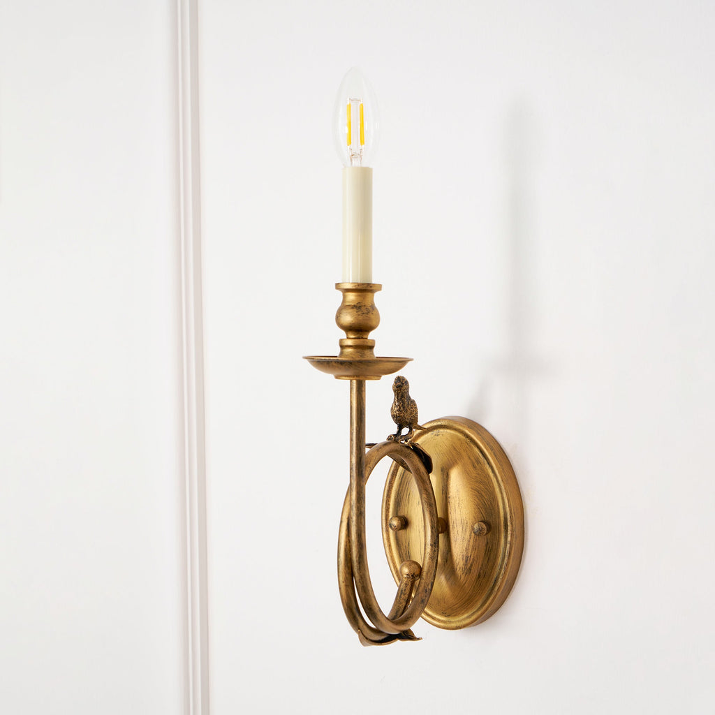 Clérachic wall sconce photographed close on a white wall, the candle bulb lit and the bird perched on the ring below.