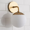 Clérachic Petal-Cap Opal Globe Wall Sconce in brass, single head, its scalloped cap set over the opal globe on white tile.