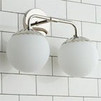 Two-head Clérachic Petal-Cap Opal Globe Wall Sconce in polished nickel, seen from below against white tile.
