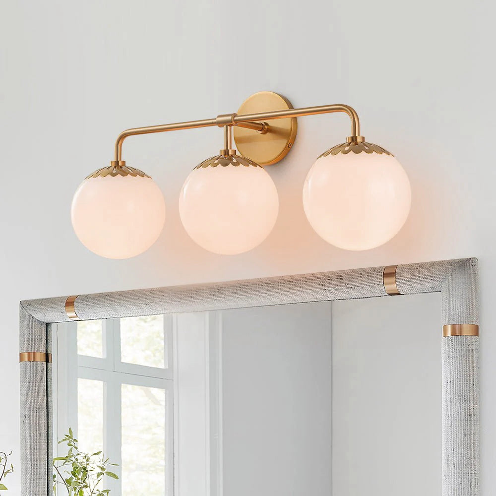 Lit brass Clérachic Petal-Cap Opal Globe Wall Sconce with three heads above a grey linen-framed mirror.