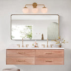 Three-head brass Clérachic Petal-Cap Opal Globe Wall Sconce lit over a mirror above an oak double vanity.