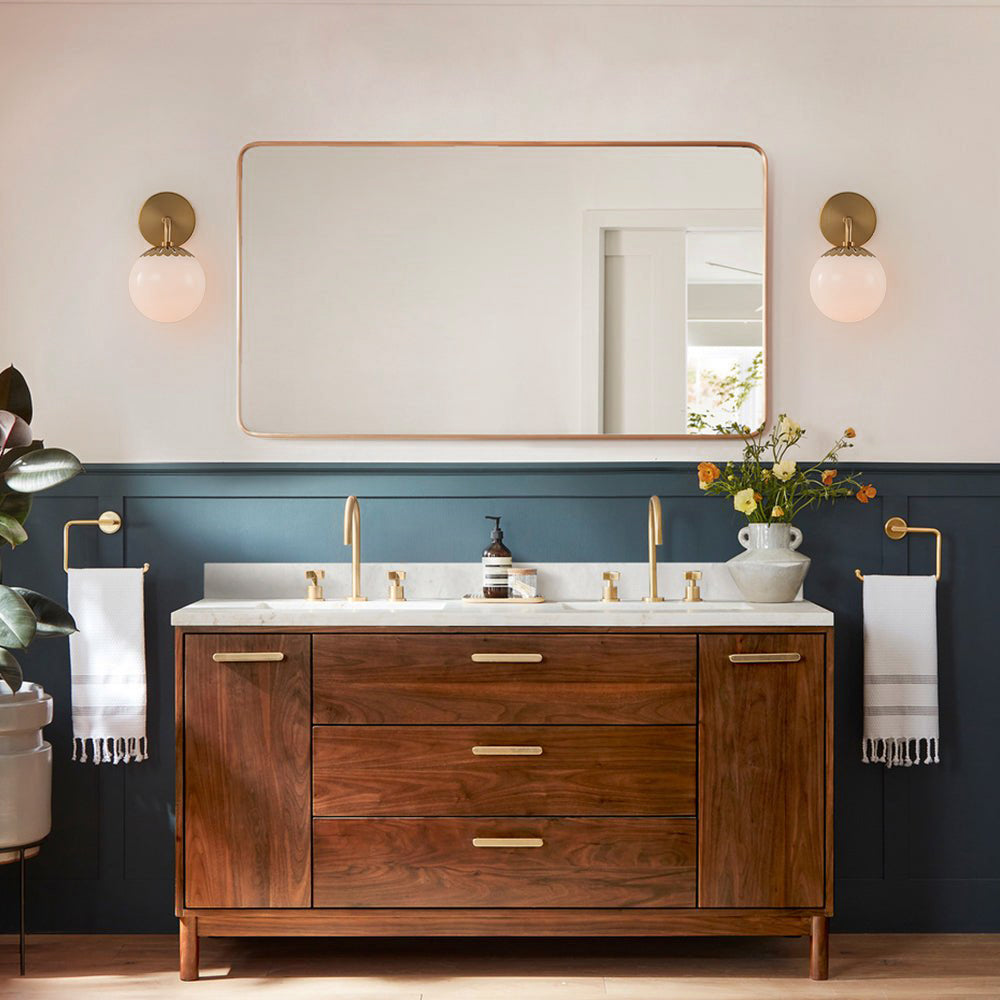 A pair of brass Clérachic Petal-Cap Opal Globe Wall Sconces flanking a mirror above a walnut vanity.