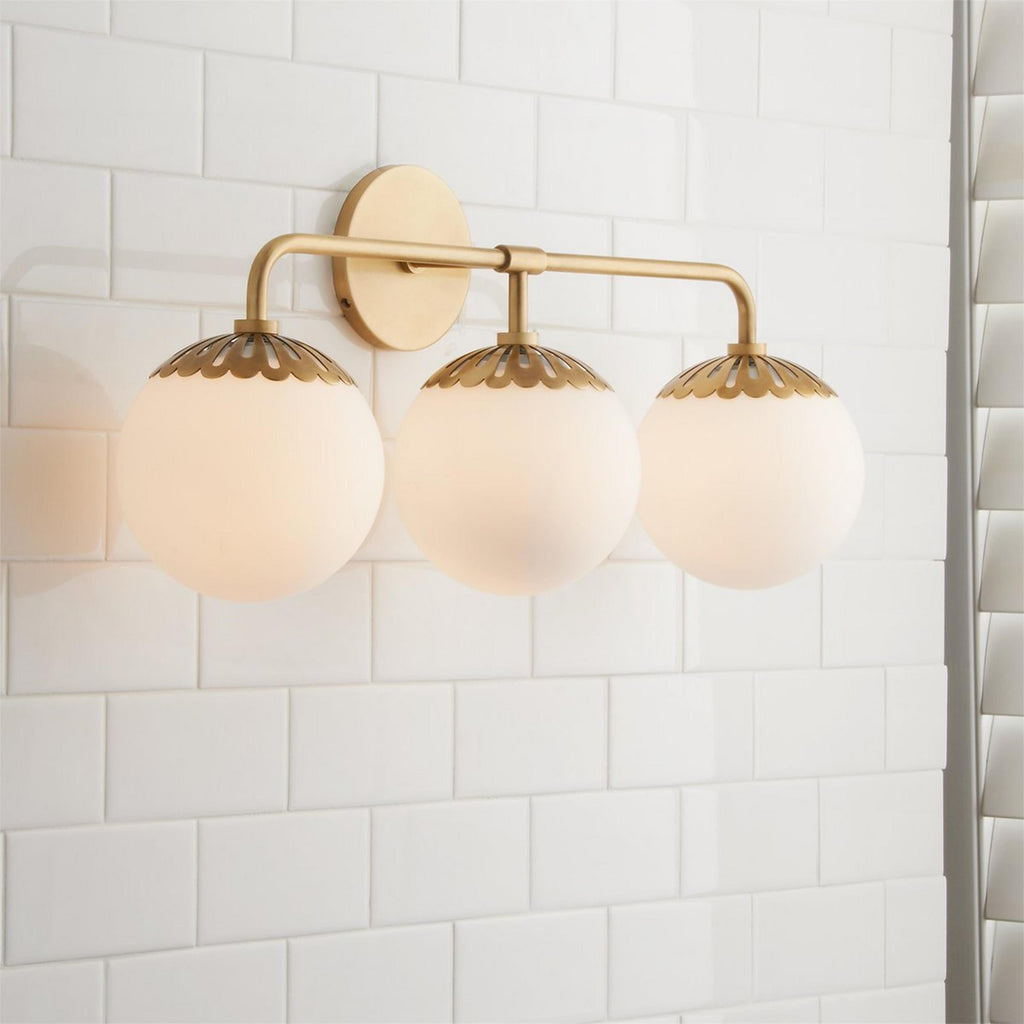 Three-head brass Clérachic Petal-Cap Opal Globe Wall Sconce angled beside a white shutter on a tiled wall.