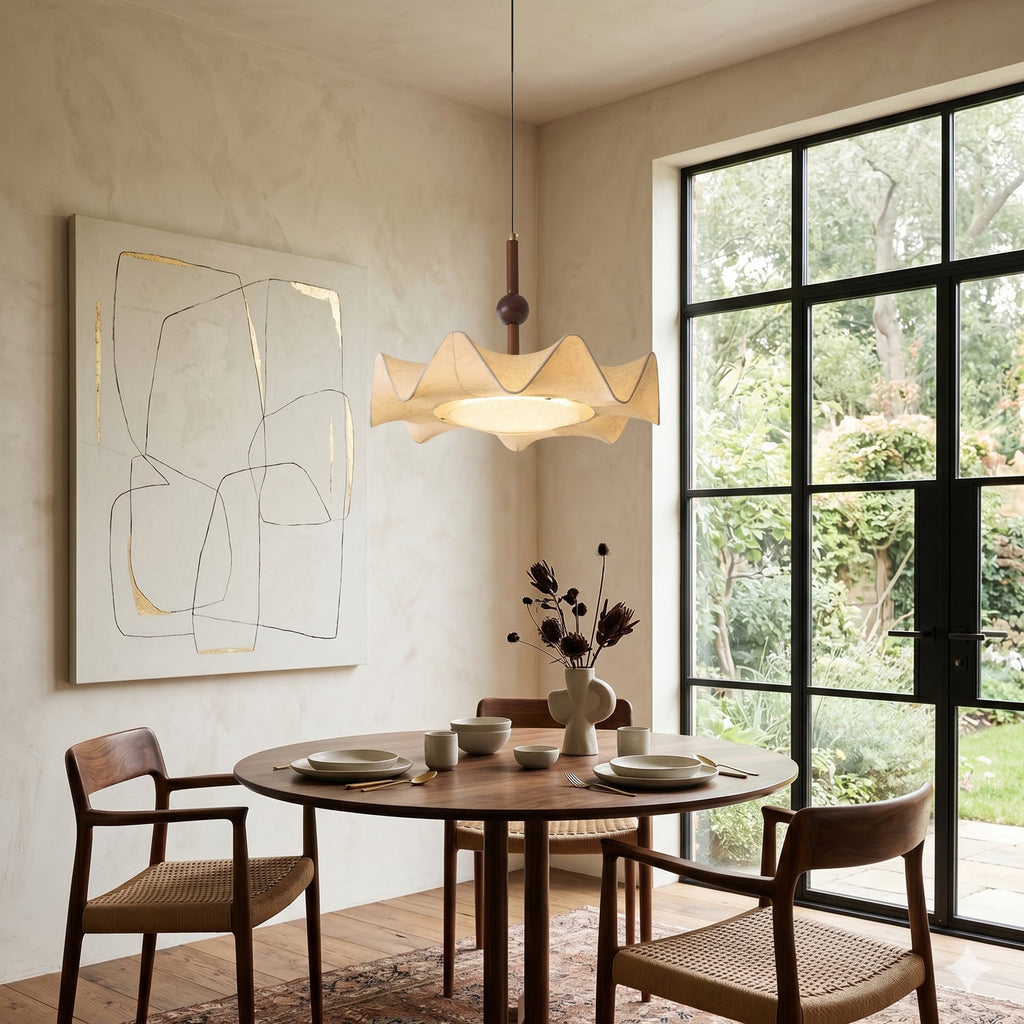A Clérachic pendant glowing warm above a round walnut dining table, beside a black-framed glazed door that opens onto a garden.