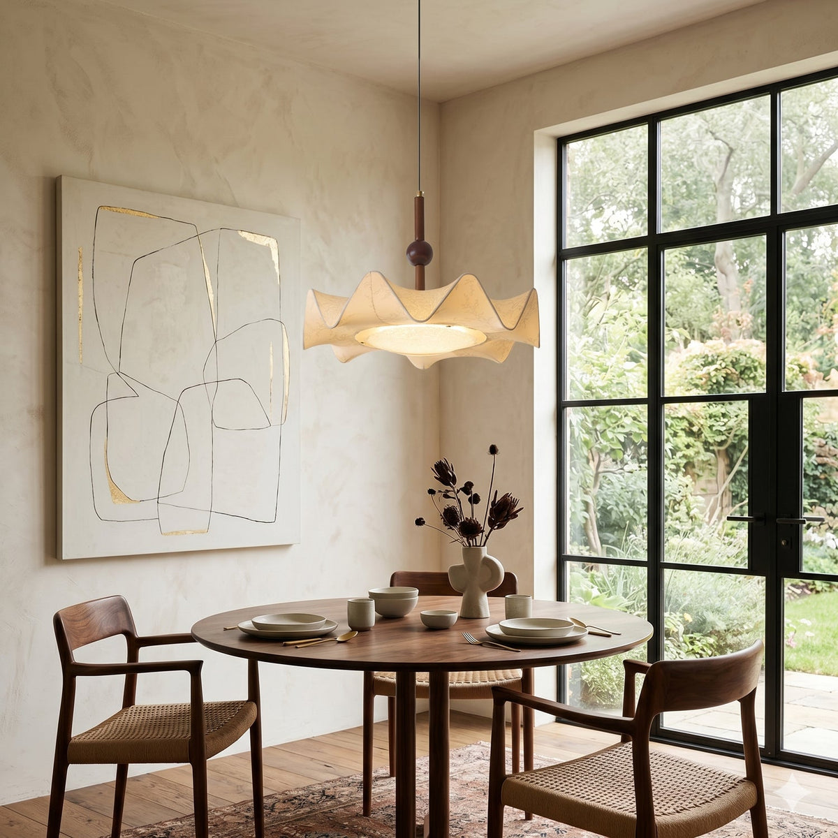 A Clérachic pendant glowing warm above a round walnut dining table, beside a black-framed glazed door that opens onto a garden.