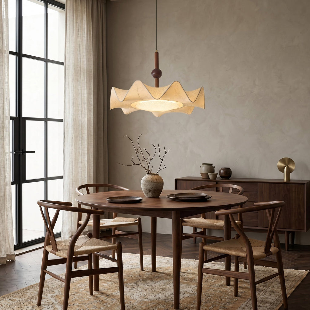 Pleated shade of this Clérachic pendant lit above a round wooden table with woven-seat chairs, a sideboard and dried branches in a vase.