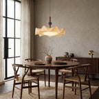Pleated shade of this Clérachic pendant lit above a round wooden table with woven-seat chairs, a sideboard and dried branches in a vase.
