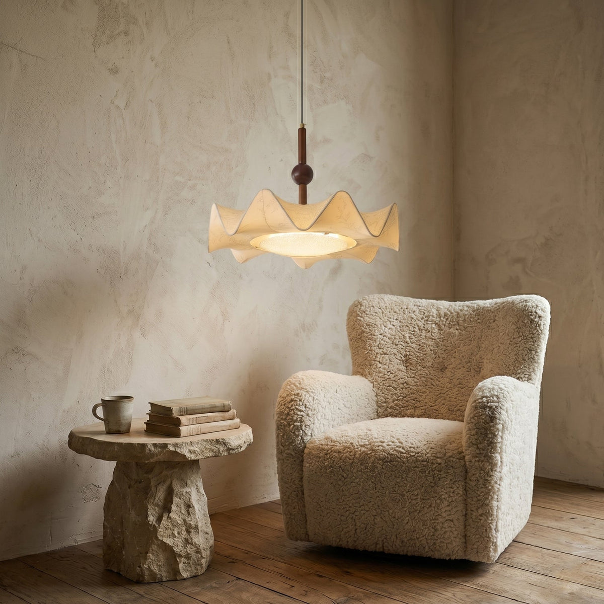 A lit Clérachic pendant on a plaster wall above a boucle armchair and a rough stone side table stacked with books and a mug.