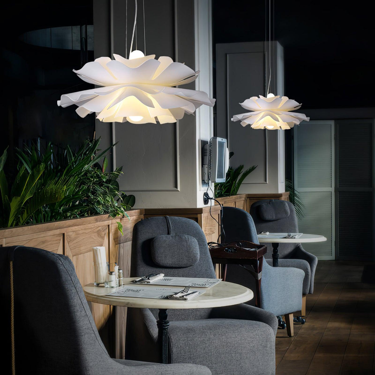 Clérachic petal pendants lit in a pair above grey tub armchairs and marble café tables among indoor planting.