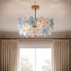 Clérachic ceiling light close up, with clear and blue glass flowers on brass arms and a stem, against a cornice and sash window.