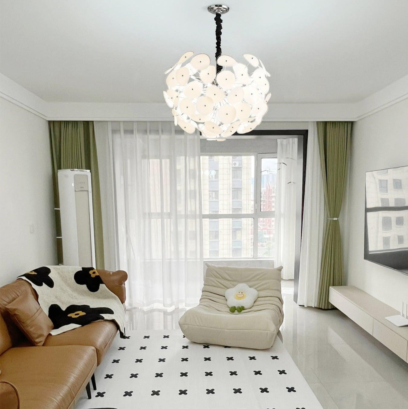 Lit Clérachic Petal Sphere Chandelier above a round glass coffee table, sheer curtains and a tall potted plant.