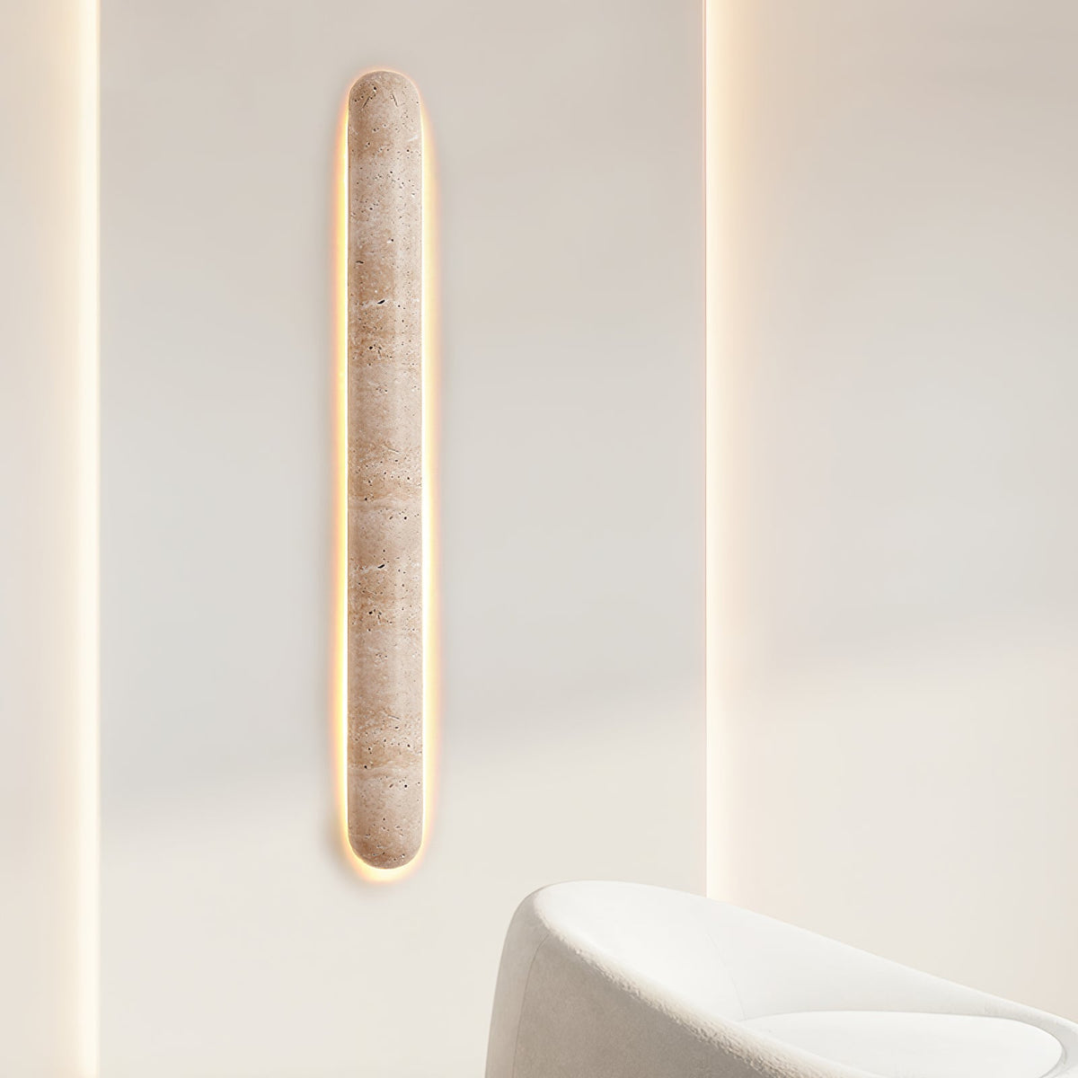 Tall pill-shaped Clérachic wall light glowing on a cream wall, with the curve of a white armchair at the lower right.
