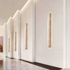 Row of Clérachic travertine wall lights set into stepped white panels along a hallway with a polished marble floor.