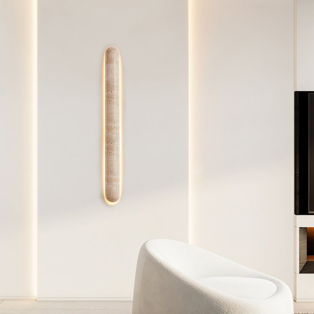 Clérachic travertine wall light lit on a white wall, with a white armchair and a dark screen at the edge of the frame.