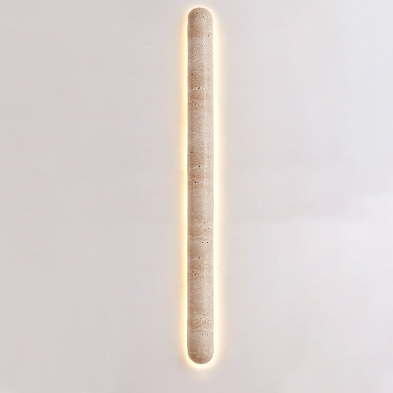 Slim, tall Clérachic wall light lit on a pale wall, its long travertine body edged with a thin warm glow.