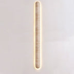 Slim, tall Clérachic wall light lit on a pale wall, its long travertine body edged with a thin warm glow.