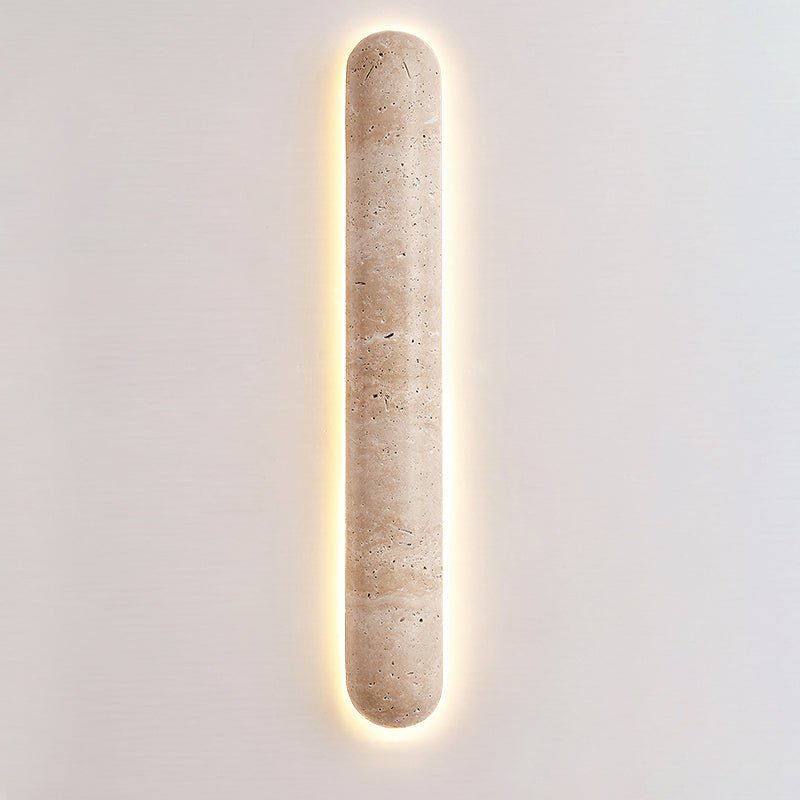 Clérachic travertine wall light with a wide warm halo, its pill-shaped body centred on a light grey wall.