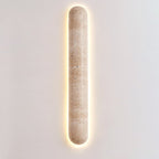 Clérachic travertine wall light with a wide warm halo, its pill-shaped body centred on a light grey wall.