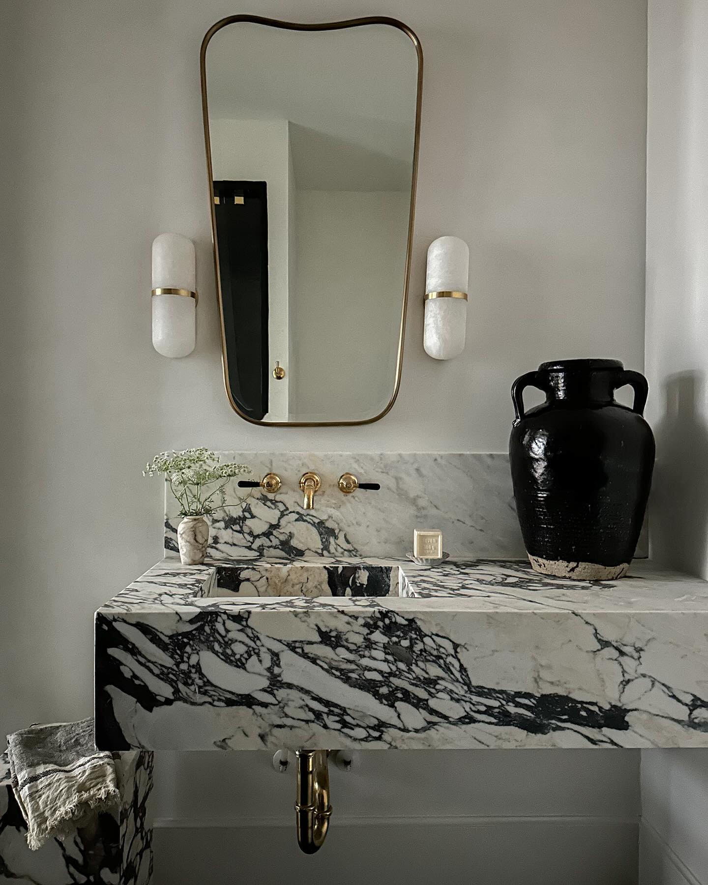 A pair of Clérachic Pill-Shaped Wall Sconce fittings flanking a brass mirror above a veined marble washbasin.