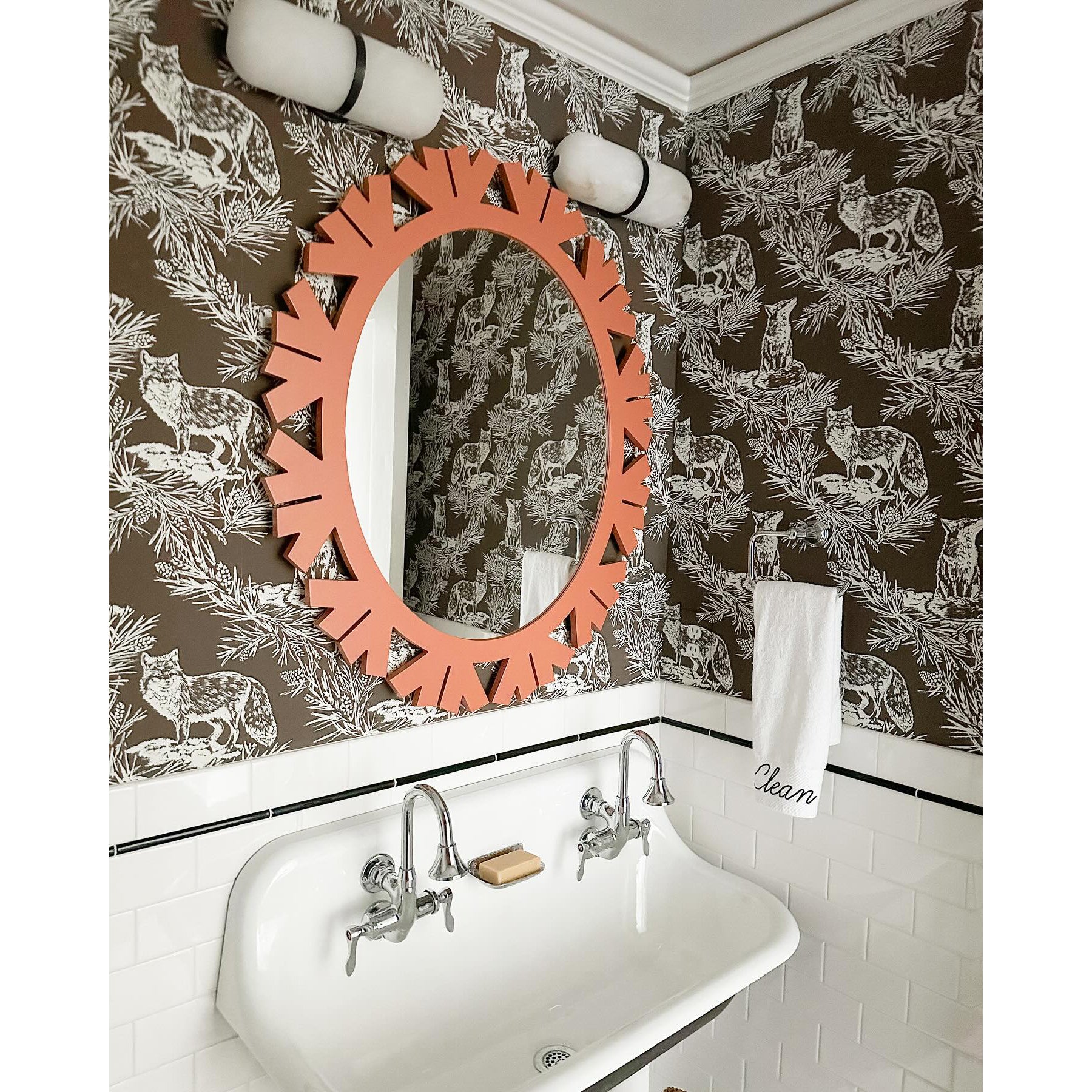 Two Clérachic Pill-Shaped Wall Sconce units mounted sideways as low bars above a coral oval mirror in a papered cloakroom.