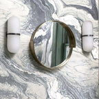 Clérachic Pill-Shaped Wall Sconce pair unlit on a grey marble wall, hung level with a round brass-framed mirror between them.