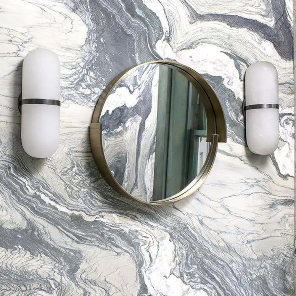 Clérachic Pill-Shaped Wall Sconce pair unlit on a grey marble wall, hung level with a round brass-framed mirror between them.