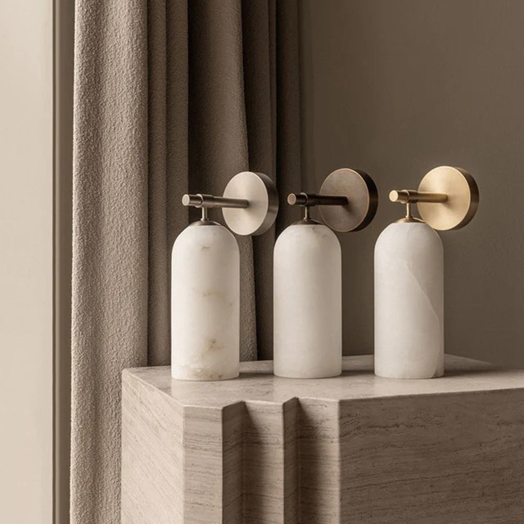 Three Clérachic Pillar wall lights grouped on a travertine plinth in front of linen curtains.
