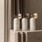 Three Clérachic Pillar wall lights grouped on a travertine plinth in front of linen curtains.